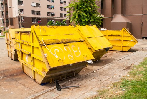 A large yellow metal skip bin with four sides folded inward, placed on a paved urban surface near a grassy area, used for rubbish removal in an apartment complex or residential block. The skip exhibits visible signs of weathering, with rust marks and grime on its surface, and has partially open panels at the top. In the background, there are multi-storey residential buildings with windows and balconies, and a bright green tree providing some greenery amidst the concrete environment. The skip is positioned on the edge of a sidewalk or driveway, with the surrounding ground showing some dirt and grass patches. This scene is representative of rubbish clearance services provided by Garden Clearance Waterloo in the Waterloo area, reflecting typical urban waste disposal practices, especially for bulky refuse removal from residential or communal areas.
