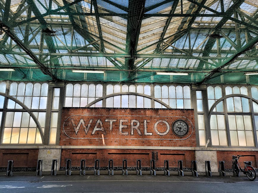 The image depicts an interior view of a transportation or railway station with a prominent brick wall displaying a large illuminated sign that reads 'WATERLOO' in capital letters, accompanied by a cir