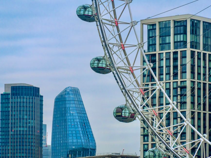 A large Ferris wheel positioned near the riverbank with multiple enclosed glass cabins evenly spaced around its circular frame, which is supported by a tall central hub and extending spokes. The wheel