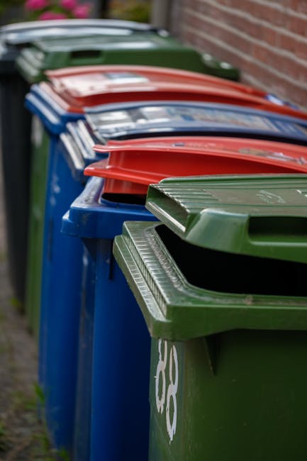 A black wheeled rubbish bin with the label 'ST. JOHN'S' positioned on the pavement alongside a street illuminated by warm streetlights. The bin is overflowing with various waste materials, including c